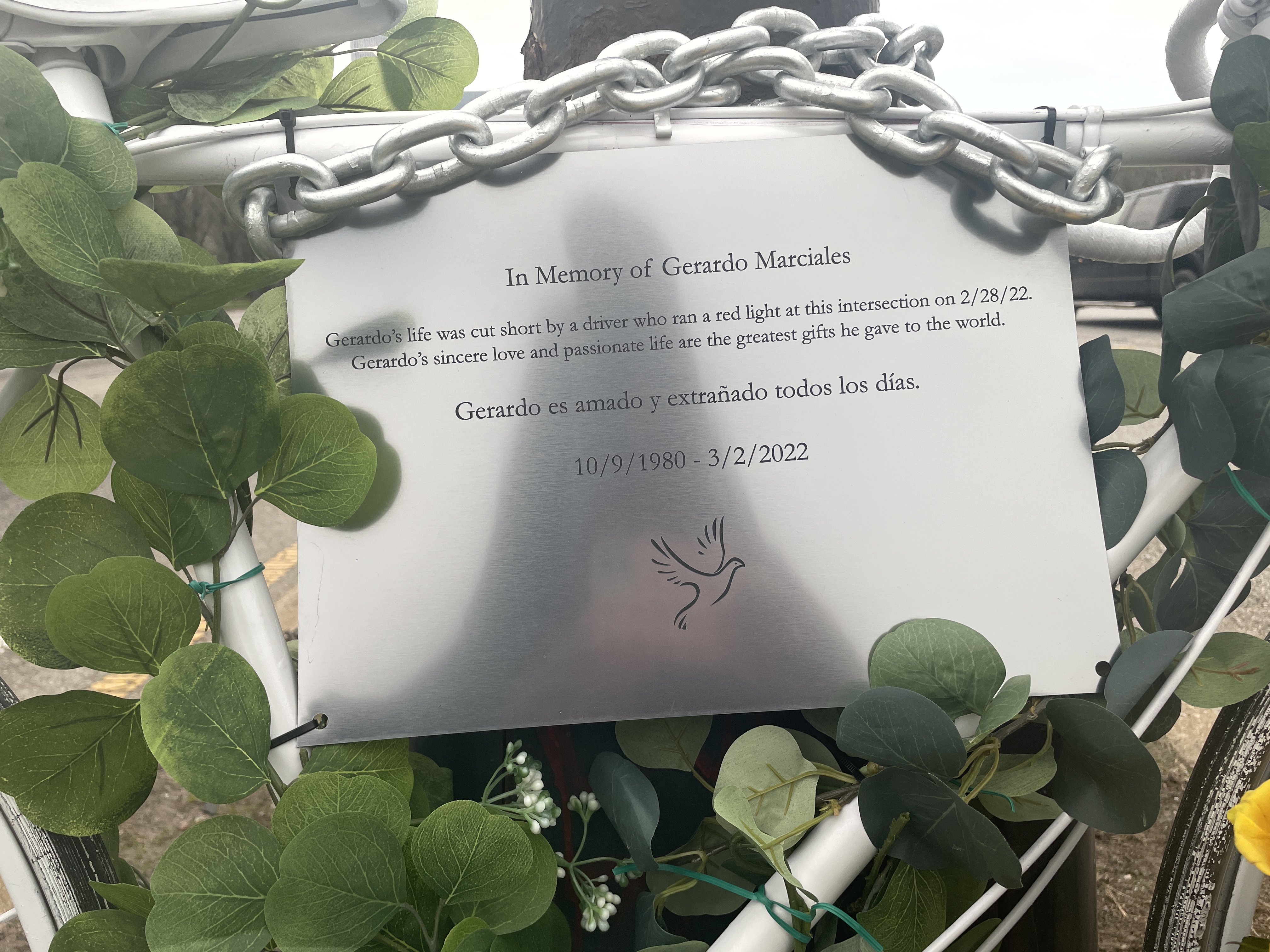 Close up of a metal placard affixed to a white ghost bike. A close-up of the placard hanging on the ghost bike in remembrance of Gerardo Marciales. It reads: "In memory of Gerardo Marciales: Gerardo‘s life was cut short by a driver who ran a red right at this intersection Gerardo sincere love and passionate life are the greatest gift he gave to the world. Gerardo es amado y extrañado todos los días. (Gerardo is loved and missed every day) 10/9/1980-3/2/2022" 
