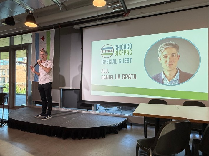 1st Ward Alderperson Daniel La Spata standing on a small stage in front of a presentation screen